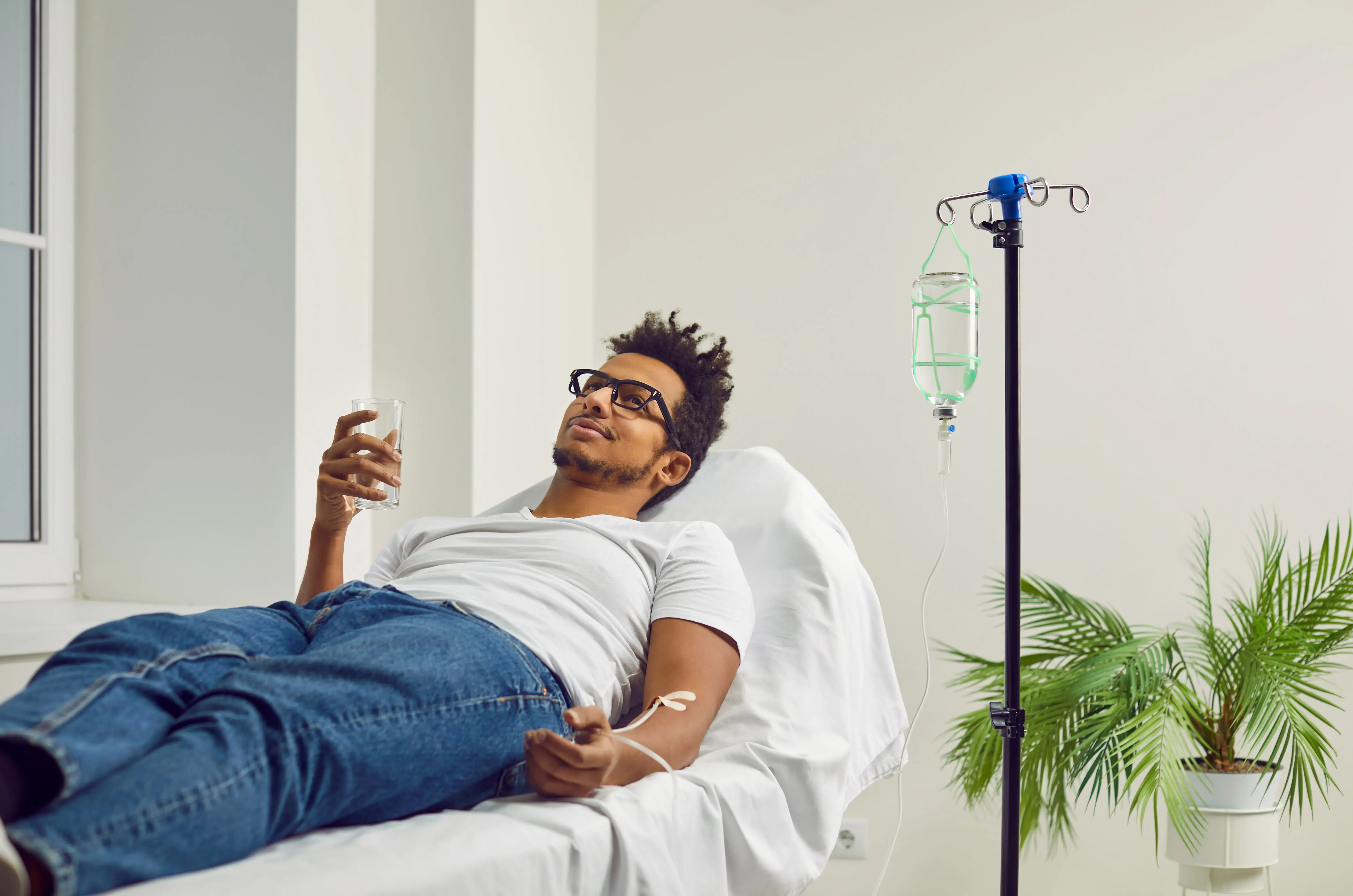 Man receiving IV vitamin infusion while relaxing on a clinic bed, holding water in a modern wellness setting
