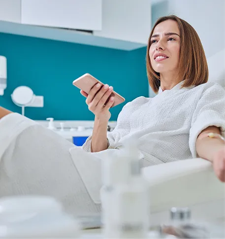 Relaxed woman in spa robe receiving IV therapy while holding smartphone in modern wellness clinic